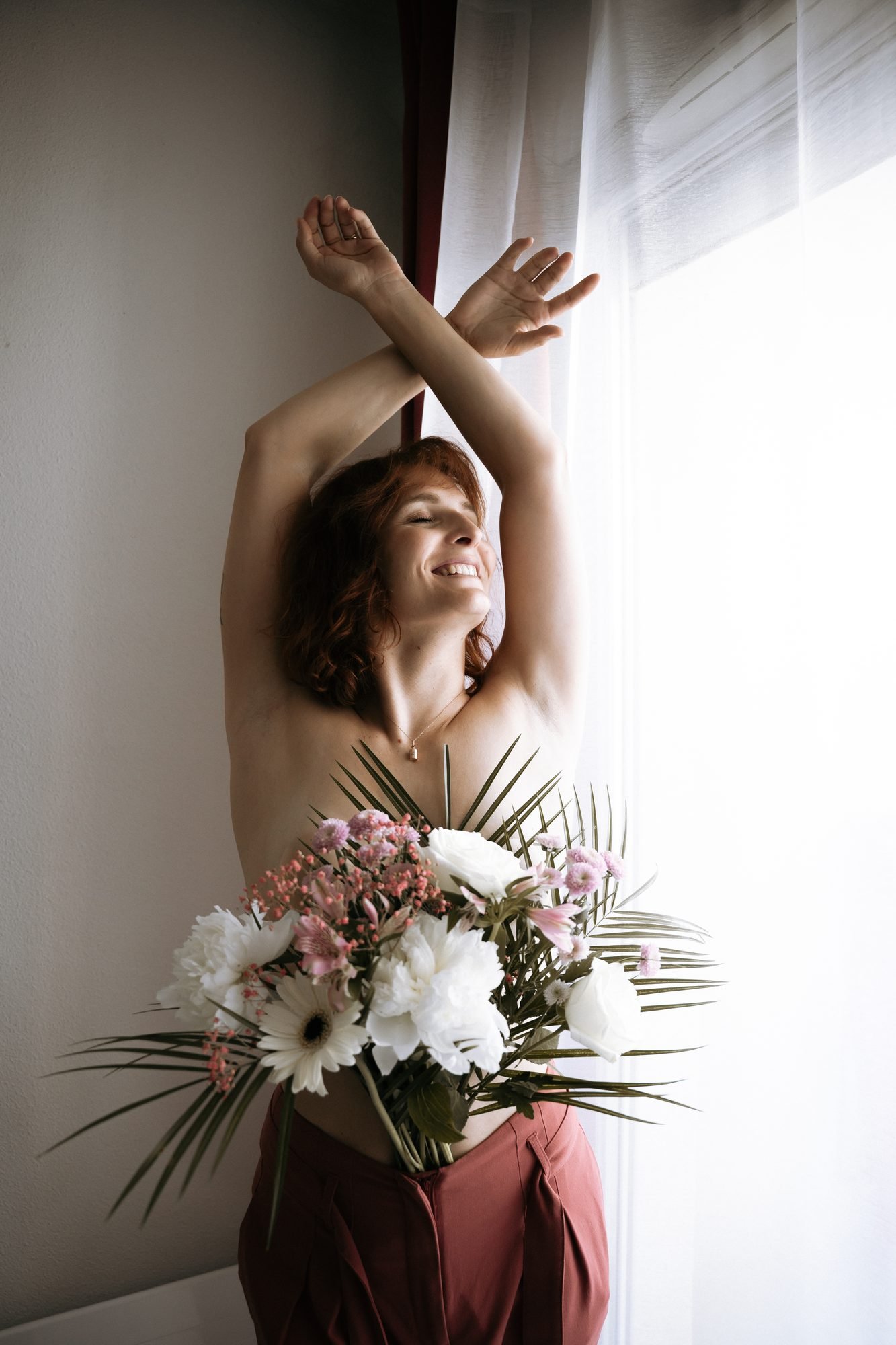 Femme souriante, bras levés, tenant un bouquet de fleurs blanches et roses près d'une fenêtre