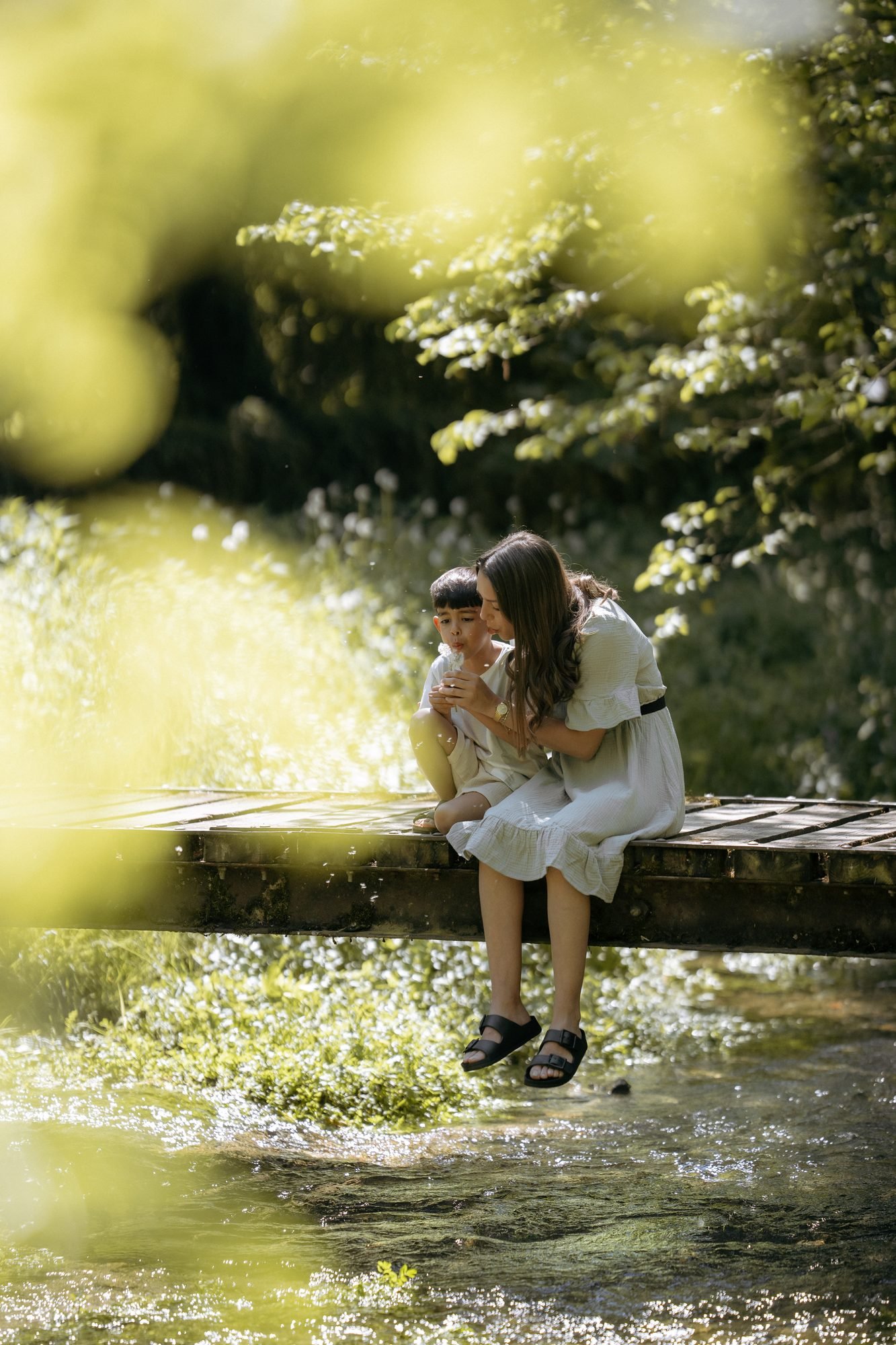 Mère et enfant assis sur un pont en bois, soufflant un pissenlit