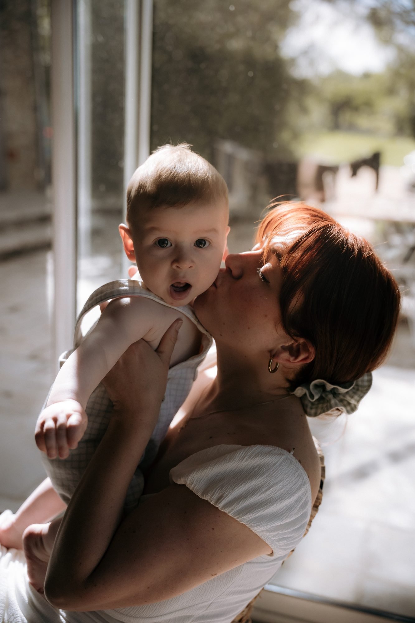 Mère embrassant son jeune enfant sur la joue, lumière naturelle près d'une baie vitrée