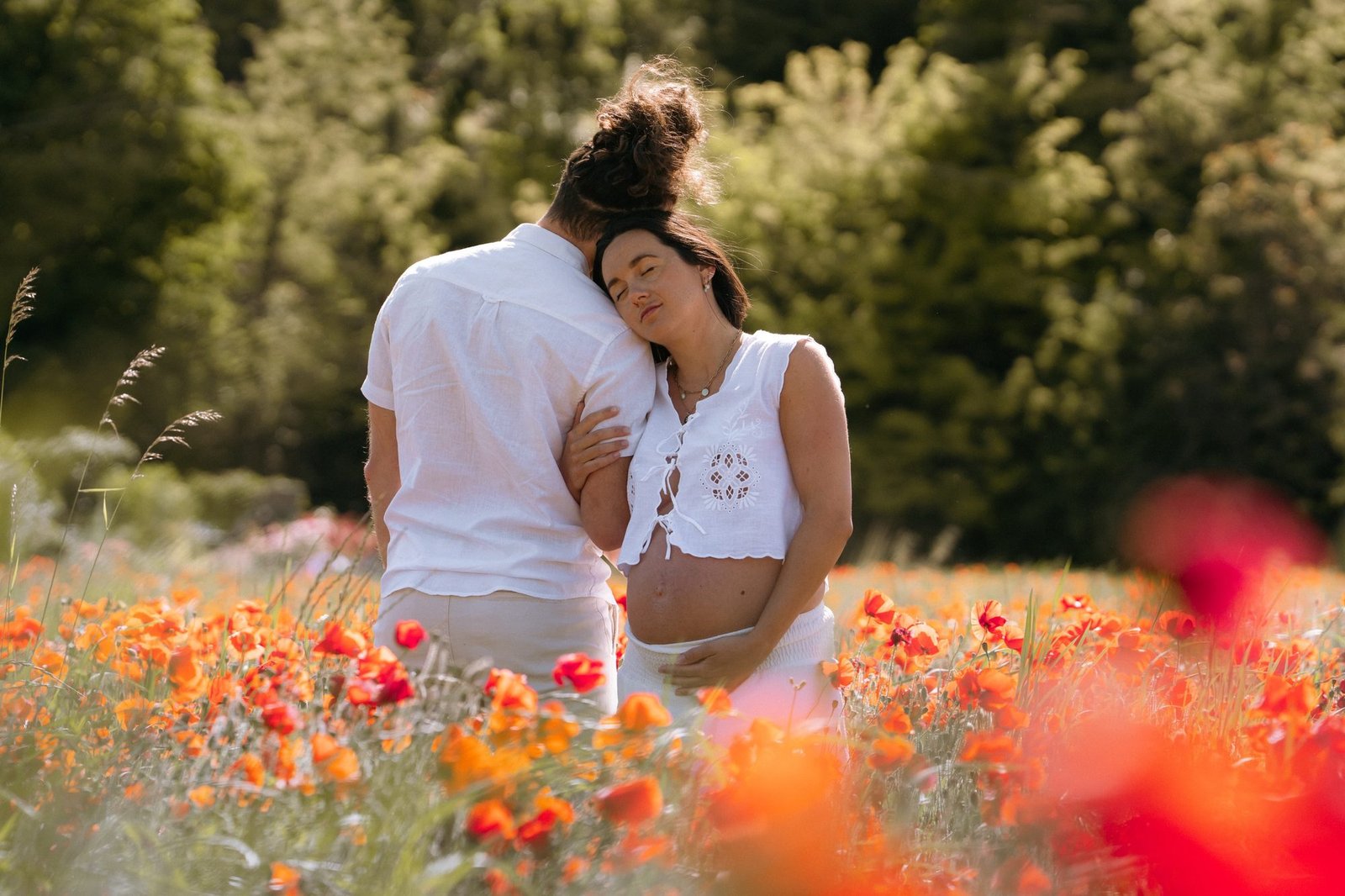 Couple enlacé dans un champ de coquelicots, femme enceinte les yeux fermés, fleurs floues au premier plan