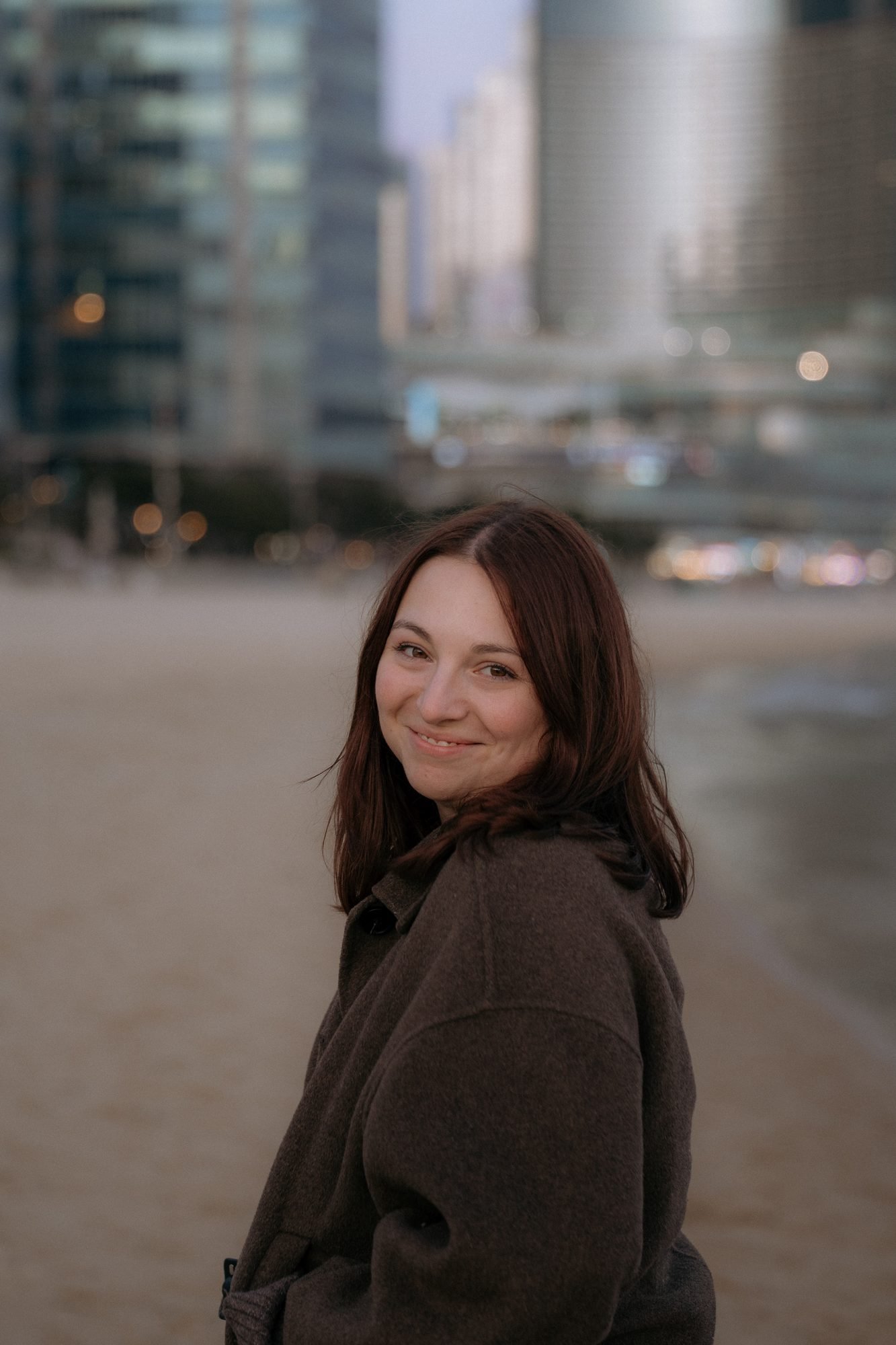 Portrait de Laetitia, photographe, souriante, cheveux châtains, devant un paysage urbain en fin de journée